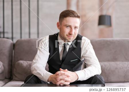A man in business clothes sits at a table with folded arms in a modern interior with soft lighting. A focused look and professional style emphasize the atmosphere of confidence and professionalism. 126895737