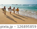 Four women walk on sandy beach holding surfboards. Female surfers silhouettes cast long shadows on sand. Ocean waves break gently nearby. Clear sky and horizon. Beach vibe with water sport activity. 126895859
