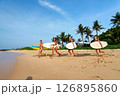 Four diverse female surfers walk with surfboards on tropical beach. Friends enjoy sunny day, palm trees. Women athletes ready for ocean waves. Active lifestyle, summer adventure. 126895860