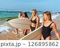 Women in swimsuits carry surfboards on sunny beach. Surfing tour. Friends ready for adventure on ocean waves. Enjoying travel, active lifestyle, tropical day. Smiling faces share joy. 126895862