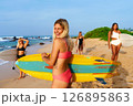 Women surfers hold surfboards on sunny beach. Friends in swimsuits enjoy coastal activities, summer vibes, ocean waves. Smiling women connect with nature, learn surfing basics at camp. 126895863