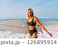 Blonde woman in black bikini with surfboard on sandy beach. Smiling surfer stands by ocean waves. Summer water sport activity, surfing adventure. Holiday at tropical sea coast. Sunny day. 126895914