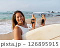 Smiling woman on sandy beach holds surfboard. Friends in swimsuits behind prepare for surfing. Ocean waves and sunny weather. Summer fun, water sports, beach vibes. 126895915