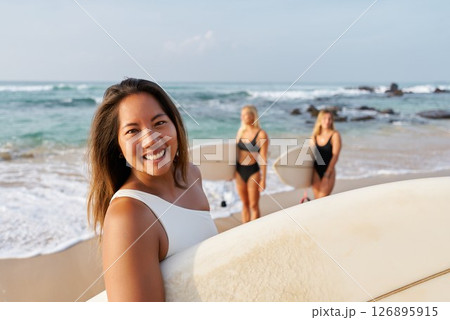 Smiling woman on sandy beach holds surfboard. Friends in swimsuits behind prepare for surfing. Ocean waves and sunny weather. Summer fun, water sports, beach vibes. 126895915
