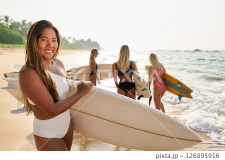 Group of young women with surfboards walk on sunny tropical beach. Surfers wear swimwear and prepare for ocean surfing. Friends enjoy water sport, adventure and travel at seaside holiday. 126895916