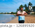 Senior man uses smart watch on tropical beach. Bearded grandpa with earbuds checks fitness tracker. Modern tech, active lifestyle, ocean background. Elderly enjoys digital innovation, relaxation. 126895920