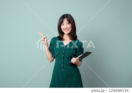 Southeast asian woman smiling while holding tablet and pointing upward, standing against plain background, representing internet technology and positive emotion Southeast asian woman smiling while holding tablet and pointing upward, standing against plain background, representing internet technology and positive emotion 126896534