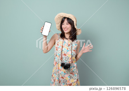 Cheerful southeast asian woman in floral dress and straw hat holding smartphone and camera, smiling against pastel green background, expressing happiness and excitement Cheerful southeast asian woman in floral dress and straw hat holding smartphone and camera, smiling against pastel green background, expressing happiness and excitement 126896578