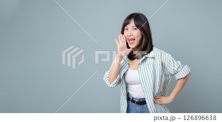 Southeast Asian woman with medium length hair wearing striped shirt and white top smiling and calling out with hand near mouth against plain background Southeast Asian woman with medium length hair wearing striped shirt and white top smiling and calling out with hand near mouth against plain background 126896638