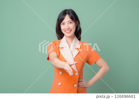 Southeast asian woman in orange suit smiling and extending hand for handshake against green background friendly gesture Southeast asian woman in orange suit smiling and extending hand for handshake against green background friendly gesture 126896681