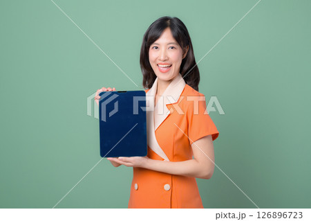 Southeast asian woman in orange suit smiling and holding digital tablet with blank screen against green background Southeast asian woman in orange suit smiling and holding digital tablet with blank screen against green background 126896723