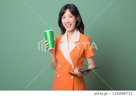 Southeast asian woman in orange dress holding green cup and tablet smiling against green background, cheerful and modern lifestyle concept 126896731
