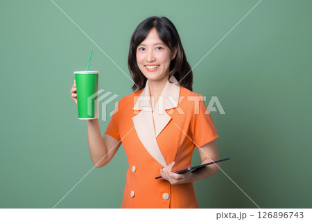 Southeast asian woman in orange dress smiling and holding green cup with straw and tablet against green background showing cheerful expression Southeast asian woman in orange dress smiling and holding green cup with straw and tablet against green background showing cheerful expression 126896743