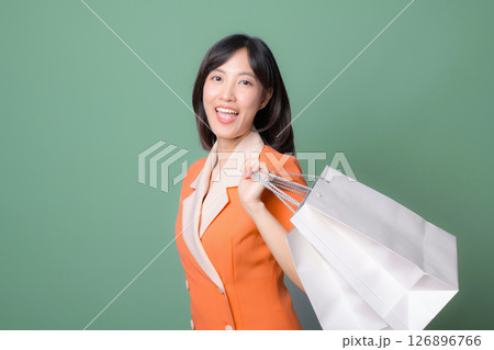 Southeast asian woman in orange blazer smiling and holding shopping bags against green background, expressing happiness and confidence in modern lifestyle 126896766