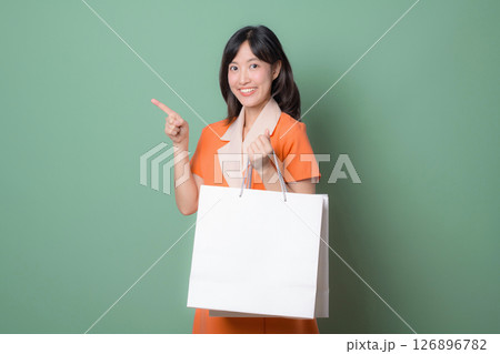 Southeast asian woman holding shopping bag smiles while pointing at something showing excitement about internet online shopping against green background 126896782