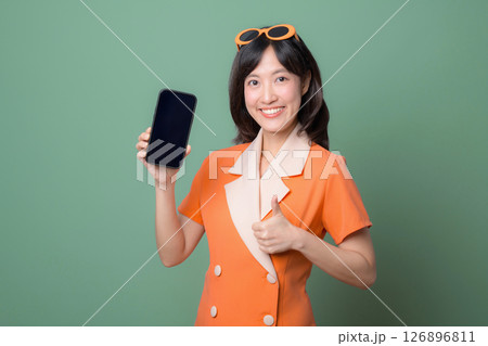 Southeast asian woman in orange dress smiling and showing smartphone with blank screen while giving thumbs up gesture on green background 126896811