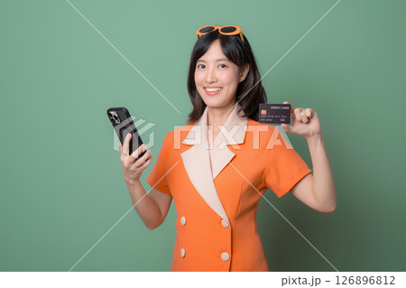Southeast asian woman in orange dress smiling holding smartphone and credit card against green background showing cheerful expression 126896812