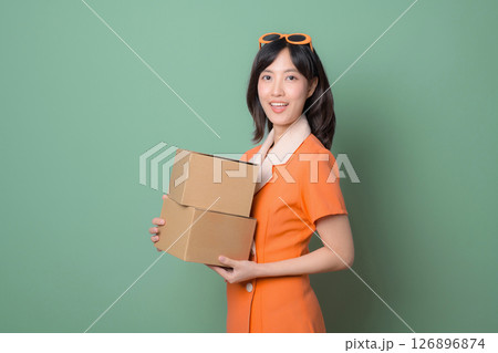 Cheerful woman in orange dress holds cardboard boxes against green background, ready for delivery or moving, expressing happiness 126896874