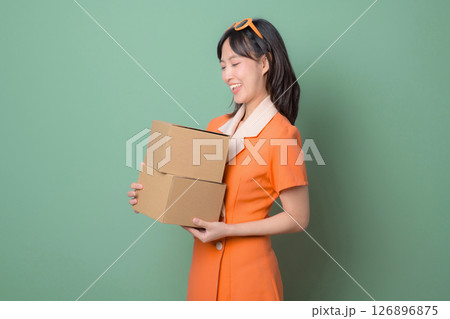 Southeast asian woman in orange dress smiling while holding two cardboard boxes against green background 126896875
