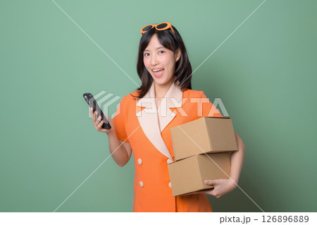 Happy woman in orange dress holds cardboard boxes and phone against green background, delivery and e commerce concept Happy woman in orange dress holds cardboard boxes and phone against green background, delivery and e commerce concept 126896889