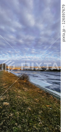 Serene sunrise over a partly frozen river with modern buildings in the background. The sky is filled with clouds, a pathway runs alongside, offering a peaceful morning scene. Serene sunrise over a partly frozen river with modern buildings in the background. The sky is filled with clouds, a pathway runs alongside, offering a peaceful morning scene. 126897679