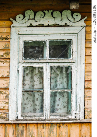 Old weathered window with carved valance showing peeling paint and floral curtains in a wooden house 126898106