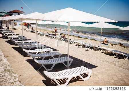 Tourists relaxing on the beach of Koktebel in Crimea with white sunbeds and umbrellas Tourists relaxing on the beach of Koktebel in Crimea with white sunbeds and umbrellas 126898168