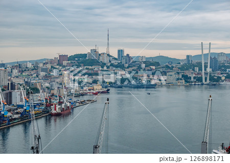 Cranes, cargo ships, and russian navy warships fill the foreground of golden horn bay, with vladivostok's skyline rising behind 126898171