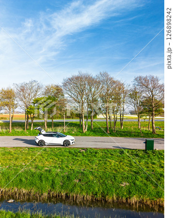 Aerial view of vibrant tulip fields in full bloom stretching along a calm canal, with farmland and green pastures under a soft morning sky in the Dutch countryside. 126898242