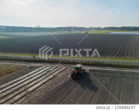 A tractor works on a large agricultural field near a road, with neat rows of soil stretching into the distance. Morning sun casts soft light across the landscape. 126898247