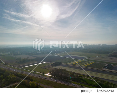 Aerial panoramic view of the Dutch countryside featuring farmland, green fields, forests, and a highway stretching through the landscape toward a distant town. 126898248