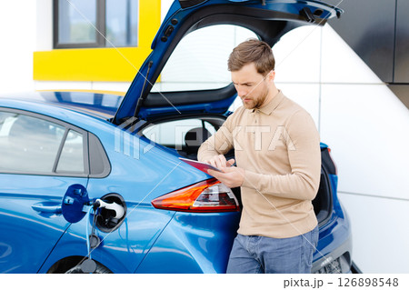 A young man is charging an electric car while standing at a station and holding a tablet A young man is charging an electric car while standing at a station and holding a tablet 126898548
