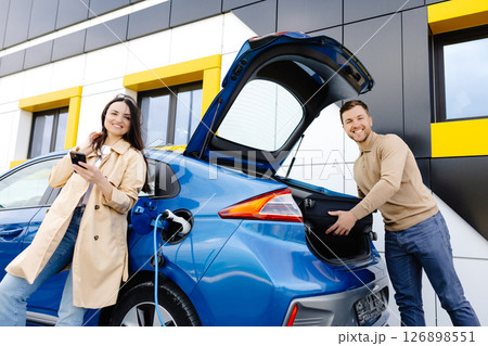 Young couple man and woman traveling by electric car having stop at charging station. The man taking a suitcase from the trunk. Man talking with girlfriend and she holding mobile phone in hand Young couple man and woman traveling by electric car having stop at charging station. The man taking a suitcase from the trunk. Man talking with girlfriend and she holding mobile phone in hand 126898551