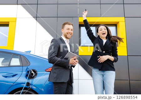 Happy female client standing at brand new car and taking keys from dealer in dealership outdoor. The woman is very happy with the new car, holding the keys in her hands Happy female client standing at brand new car and taking keys from dealer in dealership outdoor. The woman is very happy with the new car, holding the keys in her hands 126898554