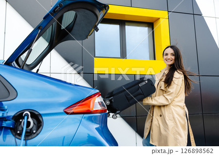 A young woman stands next to an electric car while charging the car at the station and puts her luggage in the trunk 126898562
