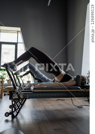 A group of woman exercises Glute bridge on a reformer bed machine in natural sunlight, focusing on pelvis and hips. A group of woman exercises Glute bridge on a reformer bed machine in natural sunlight, focusing on pelvis and hips. 126899119
