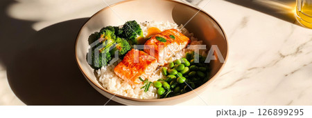 A vibrant bowl filled with salmon, rice, broccoli, and edamame is presented in a bright kitchen, showcasing a stylish and nutritious meal under warm sunlight., banner 126899295