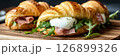 A beautifully plated croissant sandwich with a runny poached egg, savory ham, and fresh arugula, set against a bright background. A delicious and elegant morning meal with French inspiration, banner 126899326
