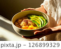 A person holds a bowl containing quinoa, sliced avocado, and fresh mango. The arrangement showcases vibrant colors, enhanced by warm sunlight 126899329