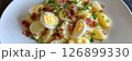 A hearty serving of Kartoffelsalat, adorned with sliced hard-boiled eggs and crispy bacon, showcases a traditional German dish served in a casual dining environment, banner 126899330