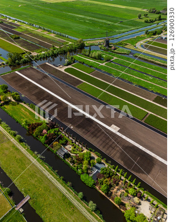 Aerial view of Dutch farmland with green fields, canals, and a traditional windmill. Neatly divided plots and vibrant vegetation highlight the beauty of rural agricultural landscape. 126900330