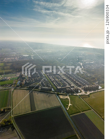 Hazy aerial view of a Dutch town merging into farmland, with the North Sea shimmering on the horizon. A mix of urban housing, fields, and forest under soft morning light. 126900334