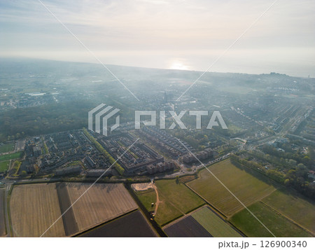Hazy aerial view of a Dutch town merging into farmland, with the North Sea shimmering on the horizon. A mix of urban housing, fields, and forest under soft morning light. Hazy aerial view of a Dutch town merging into farmland, with the North Sea shimmering on the horizon. A mix of urban housing, fields, and forest under soft morning light. 126900340