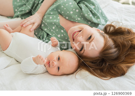 Happy family at home. Mother playing with little toddler child on bed. Mom baby relax playing having fun together. Mother looks at camera with love care. Mom of breast feeding baby rest in living room Happy family at home. Mother playing with little toddler child on bed. Mom baby relax playing having fun together. Mother looks at camera with love care. Mom of breast feeding baby rest in living room 126900369