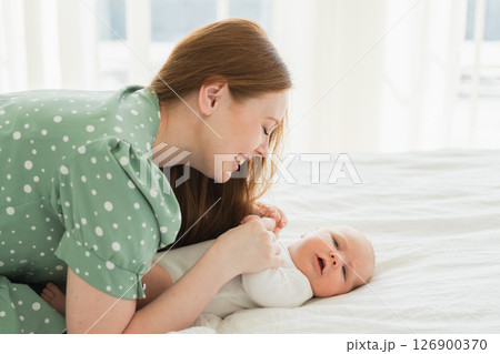 Happy family at home. Mother playing with little toddler child on bed. Mom baby relax playing having fun together. Mother looks at baby with love care. Mom of breast feeding baby rest in living room 126900370