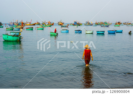 Woman in conical hat wades through coastal water surrounded by Vietnamese round fishing boats 126900804