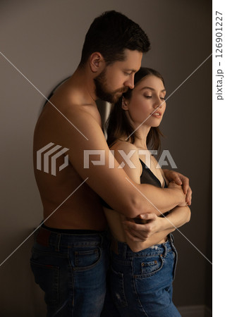 Shirtless man in jeans embracing woman in black top and jeans standing closely in intimate moment 126901227