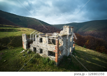 Abandoned stone building on grassy hillside, partially in ruins. Structure against backdrop of rolling hills and dense forest, under sky with dramatic clouds, evoking sense of history and solitude. Abandoned stone building on grassy hillside, partially in ruins. Structure against backdrop of rolling hills and dense forest, under sky with dramatic clouds, evoking sense of history and solitude. 126901350