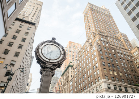 A vintage-style street clock stands prominently against a backdrop of tall, classic brick buildings in Manhattan. The upward angle and soft daylight highlight the urban charm and architectural 126901727