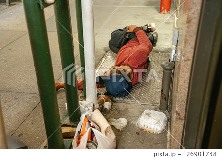 A man in a red sweatshirt sleeps on the ground beside trash on a New York street. Urban poverty and homelessness are visible in the city environment. A man in a red sweatshirt sleeps on the ground beside trash on a New York street. Urban poverty and homelessness are visible in the city environment. 126901738